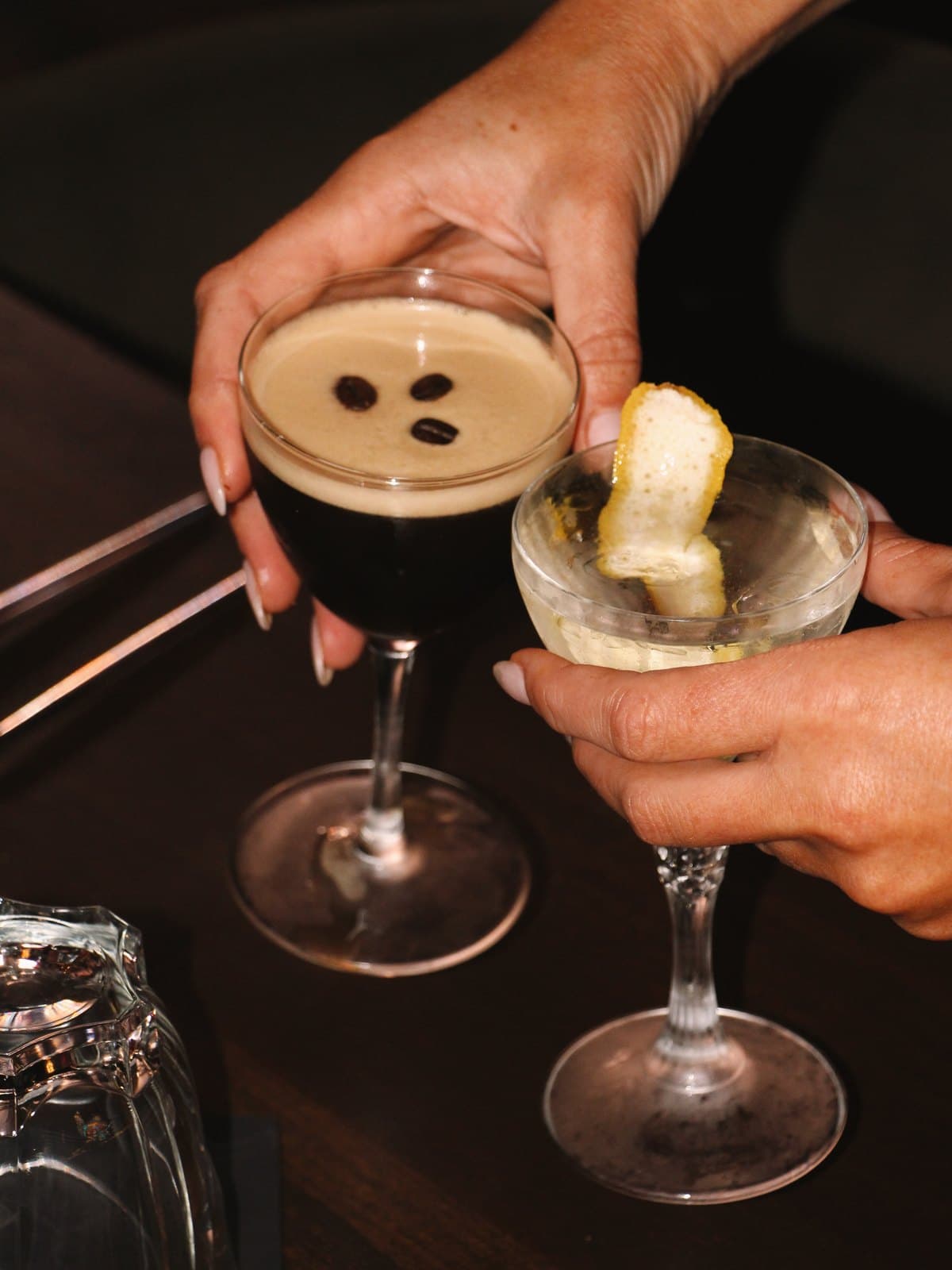 Two manicured hands raising an espresso martini and a lemon-twist martini in a toast at Jay's bar