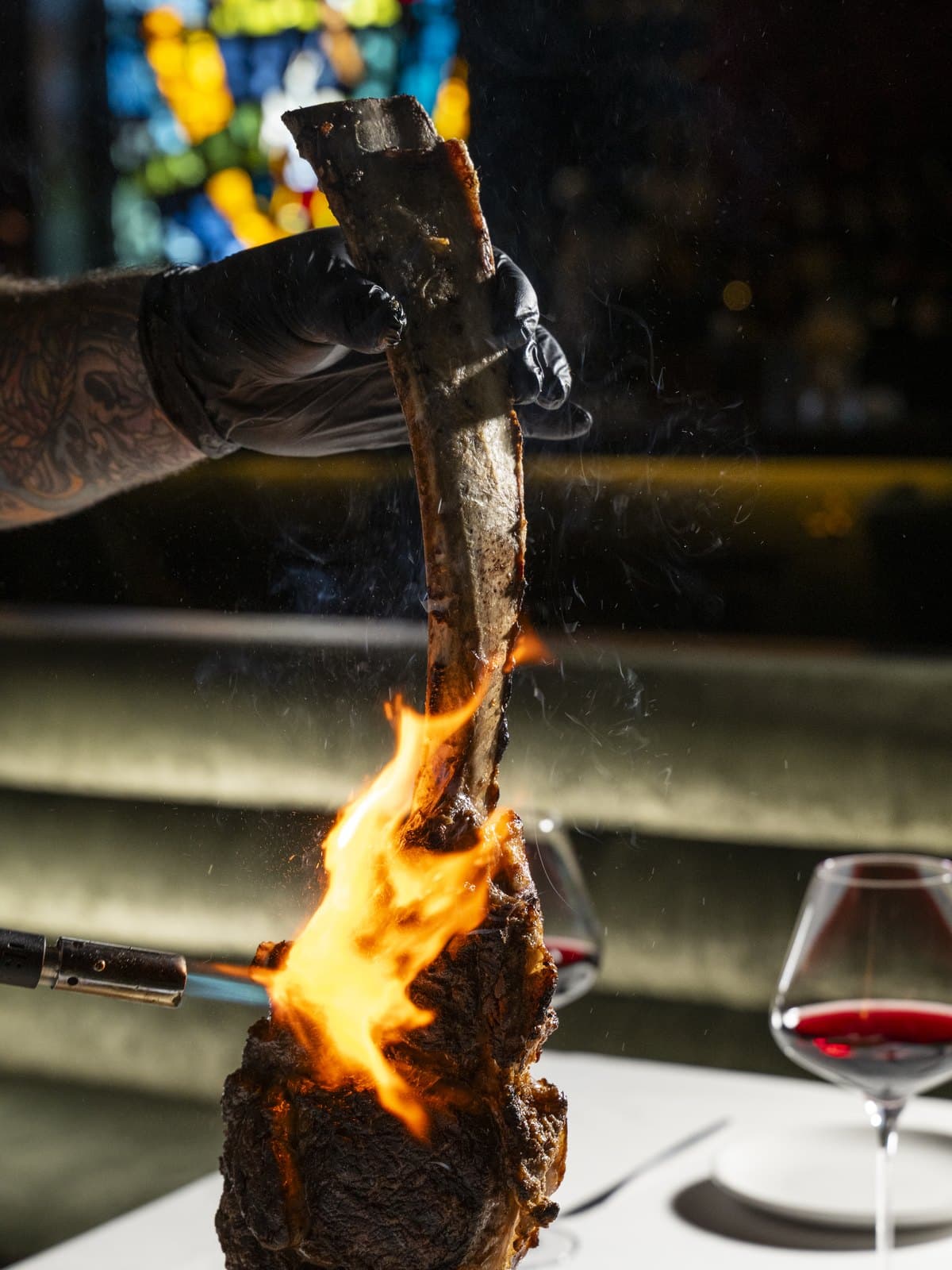 A gloved hand holds a tomahawk steak up by its long bone as a torch flames the crust, with stained glass and red wine glasses behind it at Jay's